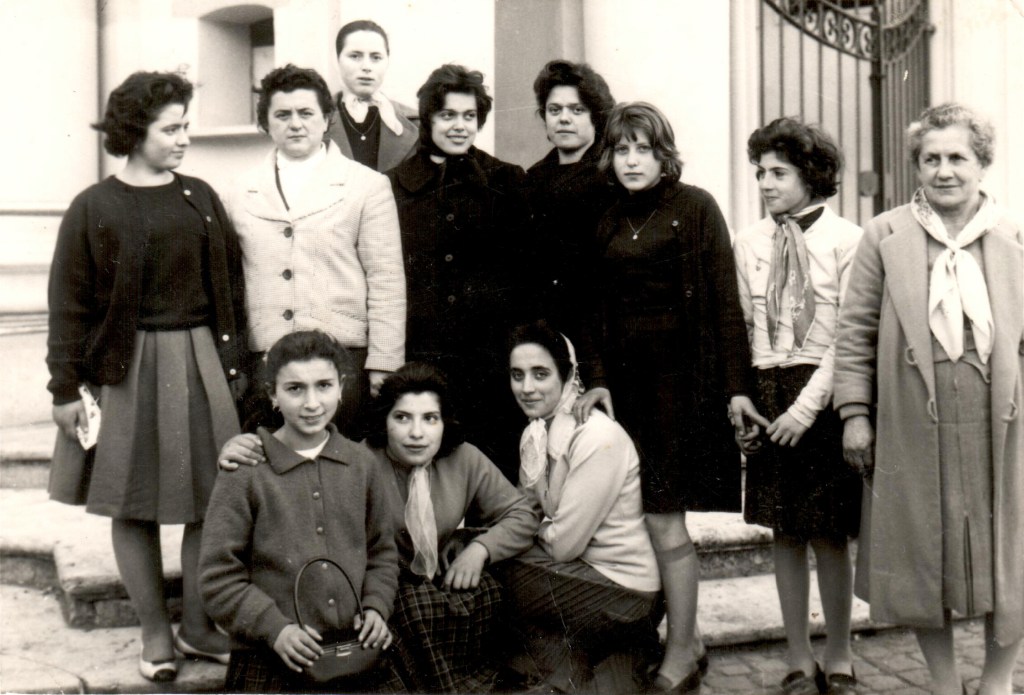 A black and white photograph of a group of women and girls standing and sitting together outside a building, smiling at the camera.