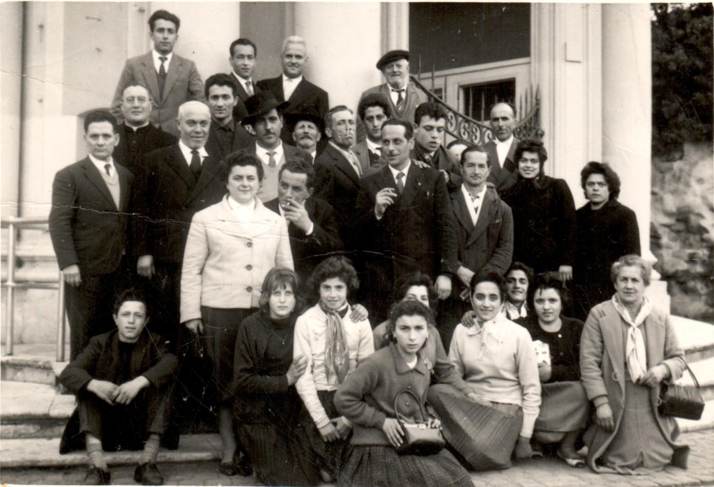 Black and white group photo of a large gathering, featuring men and women of various ages posing together outside a building.