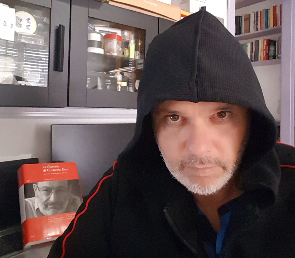 Un uomo con il cappuccio scuro e la barba corta, in primo piano, di fronte a un mobile con librerie e oggetti vari. Dietro di lui, un libro intitolato 'La filosofia di Umberto Eco' è in evidenza.