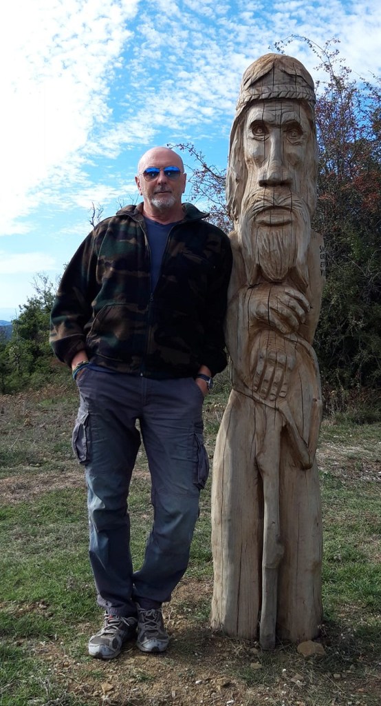 Un uomo in piedi accanto a una scultura in legno rappresentante un personaggio con barba e cappello, in un ambiente naturale sotto un cielo nuvoloso.