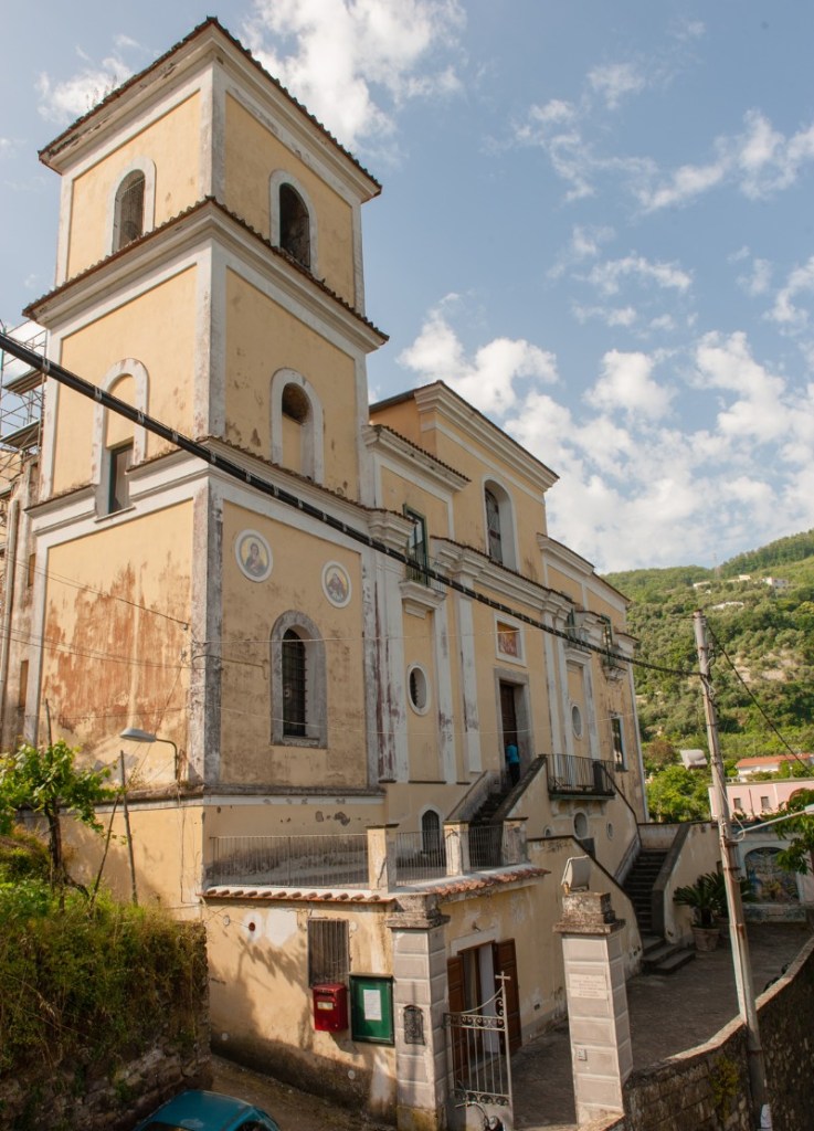 Vista esterna di un edificio religioso con pareti gialle e dettagli architettonici, situato in una zona collinare con cielo blu e nuvole.