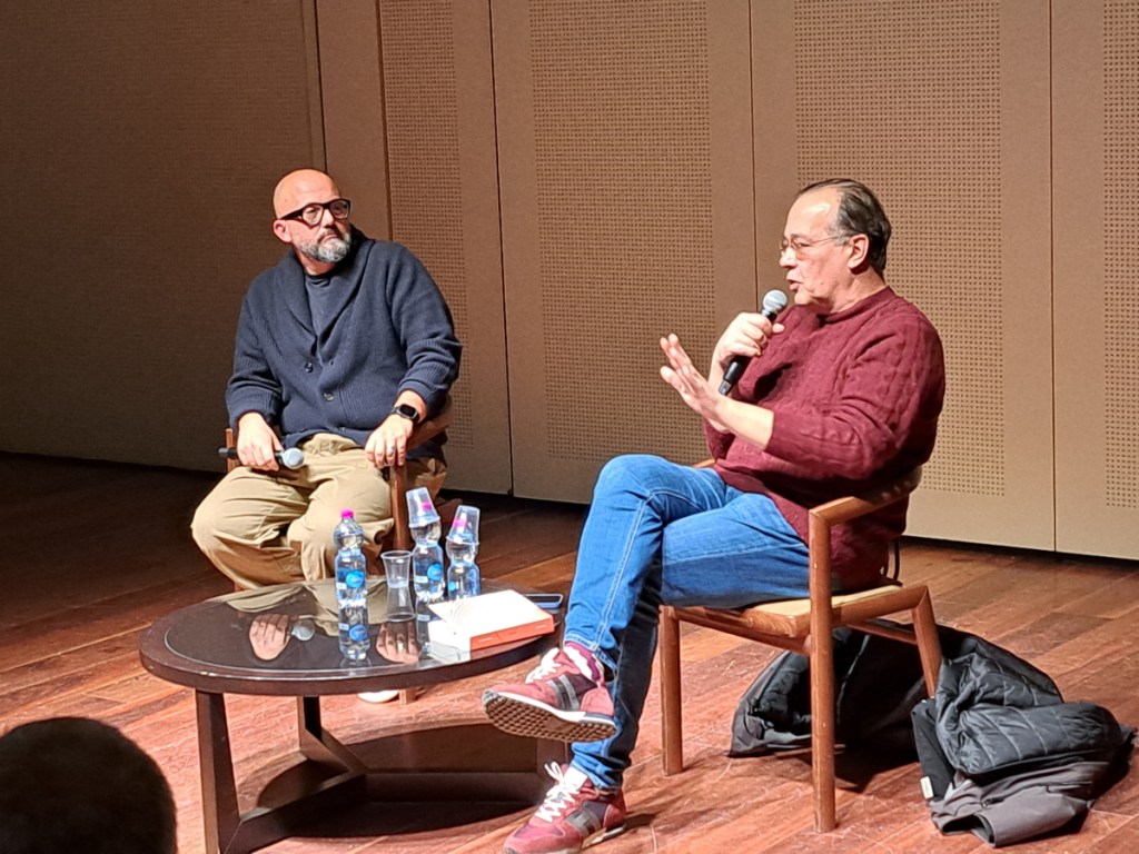 Due uomini seduti in una sala, uno dei quali parla mentre tiene un microfono. L'altro ascolta con interesse. Un tavolo con bottiglie d'acqua e un libro è visibile tra di loro.