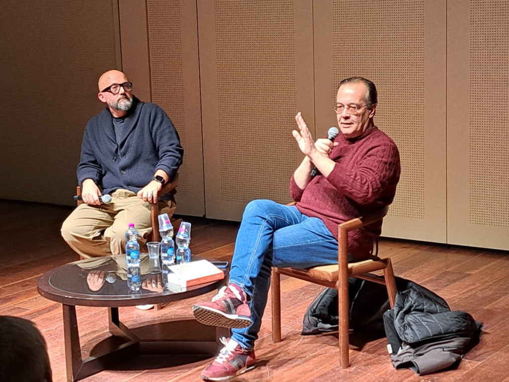Due relatori seduti su una sedia in un evento, uno con occhiali e barba corta, l'altro con maglione color bordeaux, entrambi interagendo su un tavolo con bottiglie d'acqua e libri.