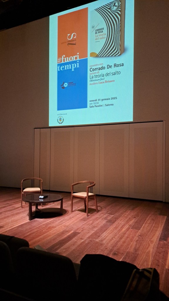 Evento di presentazione del libro 'La teoria del salto' di Corrado De Rosa, con moderatore Luca Briasco. Si vedono due sedie accanto a un tavolino su un palco palesemente preparato per un incontro.