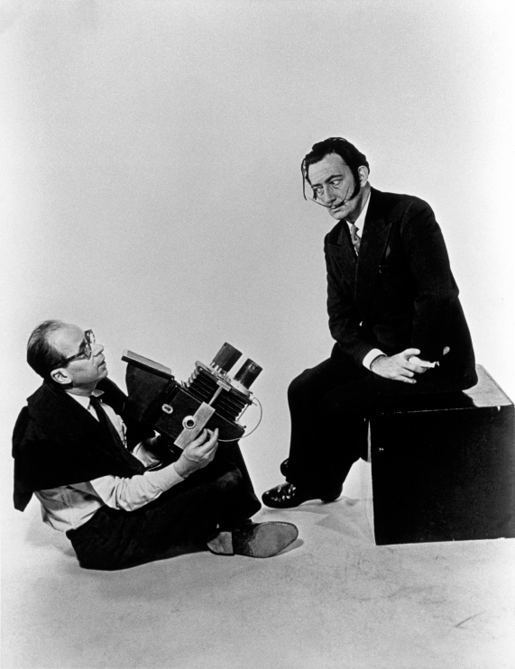 Un fotografo in posa con una macchina fotografica accanto a un uomo seduto su un cubo, in un'ambientazione minimalista.