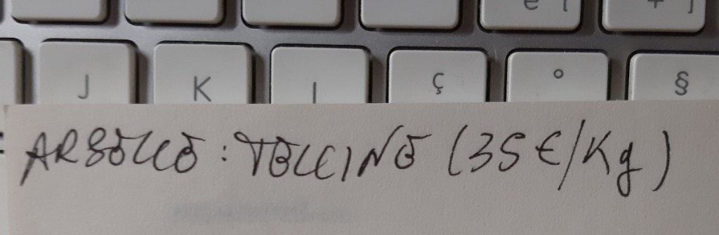 Un primo piano di una nota scritta a mano che mostra la parola 'Arbusto:' seguita da 'Tollino (35€ / Kg)' su un documento posizionato sopra una tastiera di computer.
