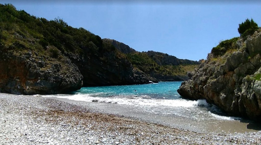Spiaggia circondata da scogli, con acque turchesi che si infrangono sulla riva sabbiosa, immersa in un'atmosfera naturale e tranquilla.