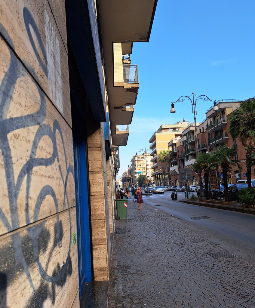 Vista di una strada con edifici residenziali e negozi, alberi e un cielo azzurro sullo sfondo.