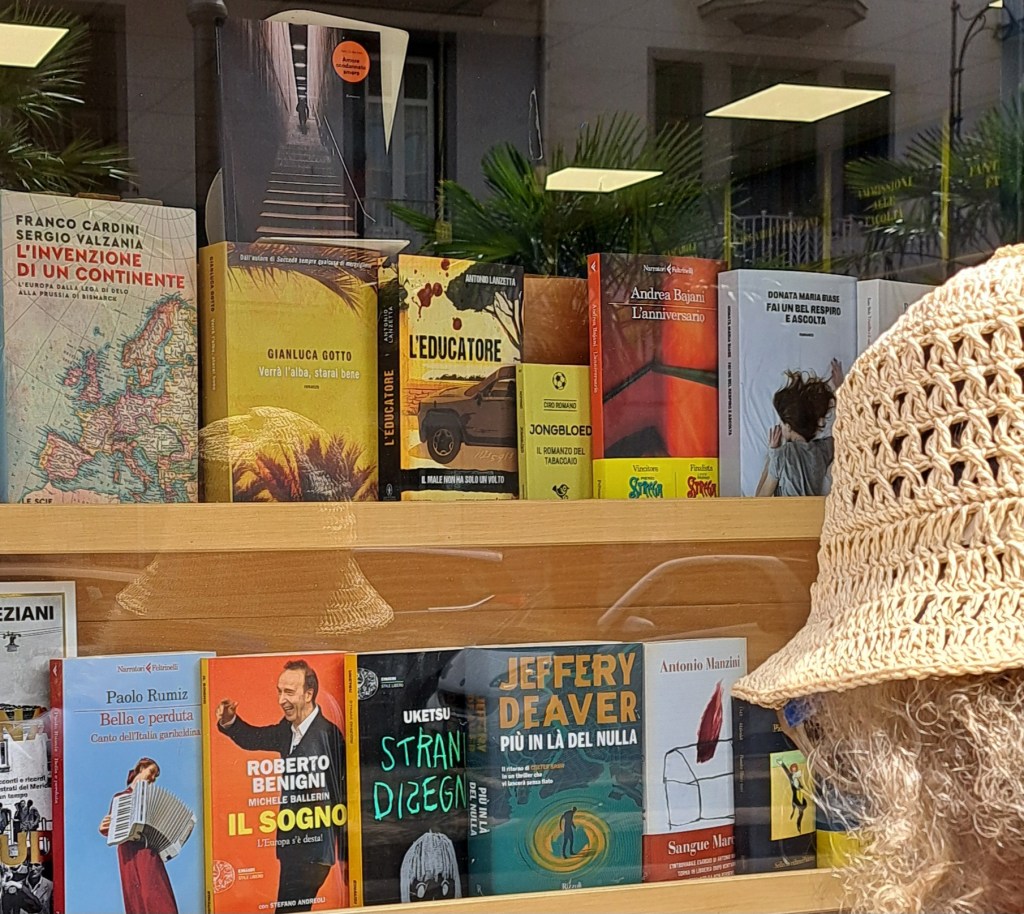 Vetrina di una libreria con una selezione di libri, inclusi titoli di vari autori e un visitatore in primo piano che indossa un cappello di paglia.