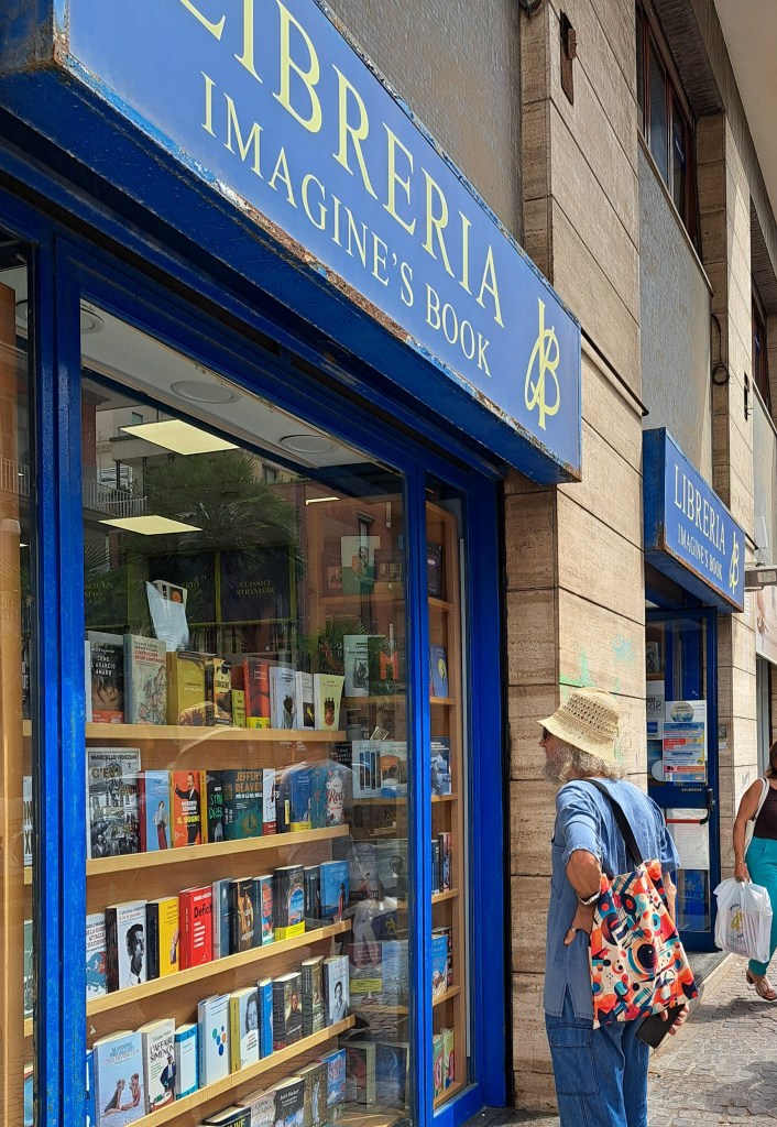 Un uomo con un cappello di paglia osserva la vetrina di una libreria chiamata 'Imagine's Book', piena di libri esposti.