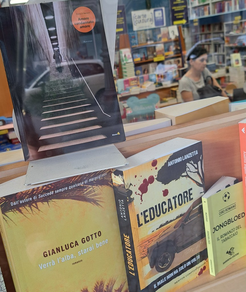 Copertina del libro 'Amore Condannato Amore' di Pietro Di Gennaro in vetrina, con riflessi e trasparenze. Sullo sfondo, si intravedono altri libri nella libreria.