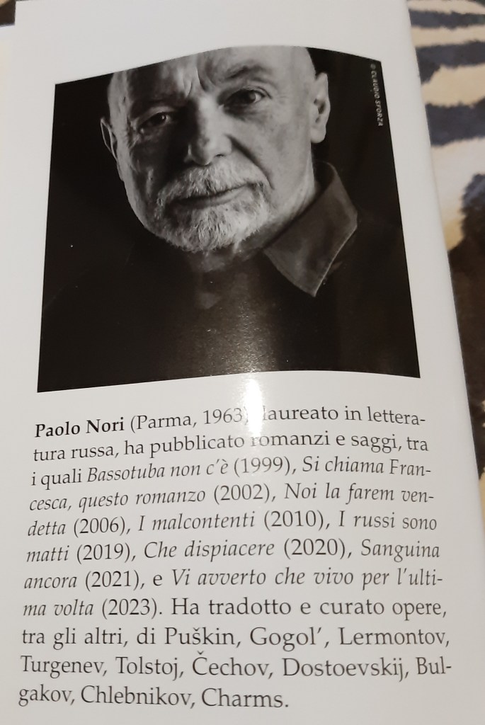 Ritratto di Paolo Nori su sfondo neutro, con espressione seria e barba bianca, mentre indossa una camicia scura.