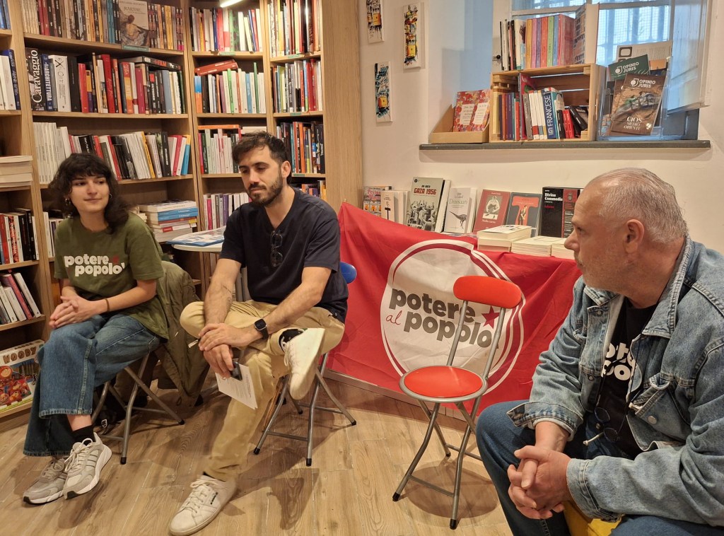Tre persone sedute in una libreria, con una bandiera rossa sullo sfondo che recita 'potere al popolo'.