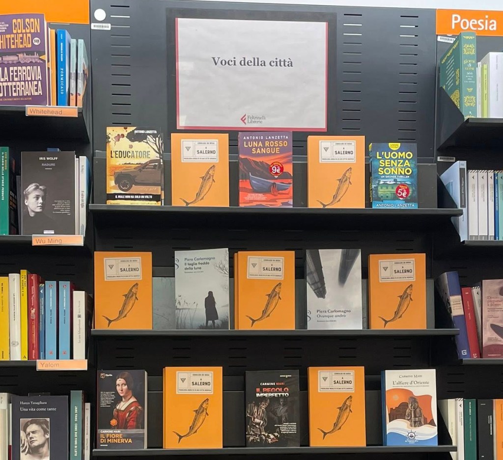 Esposizione di libri nella libreria Feltrinelli, con il segno 'Voci della città' in alto e varie copertine di libri in evidenza sugli scaffali.