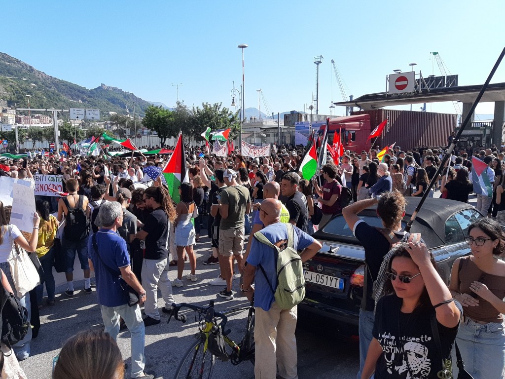 Manifestazione affollata con persone che sventolano bandiere palestinesi e cartelli, in un contesto urbano.