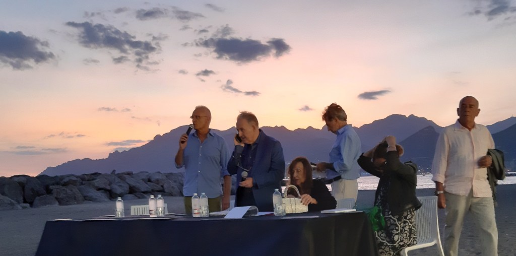 Un gruppo di persone parla e si muove durante un evento al Porticciolo di Pastena a Salerno, con un tramonto sullo sfondo.
