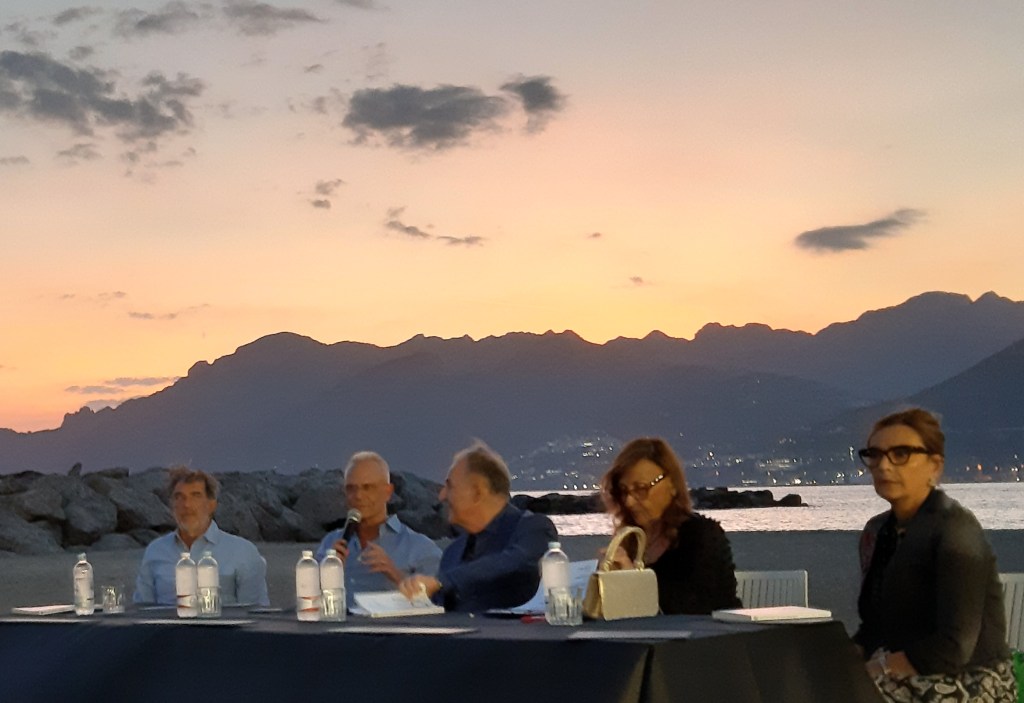 Un gruppo di relatori discute davanti a un tavolo imbottito, mentre il sole tramonta dietro le montagne su una spiaggia a Salerno.