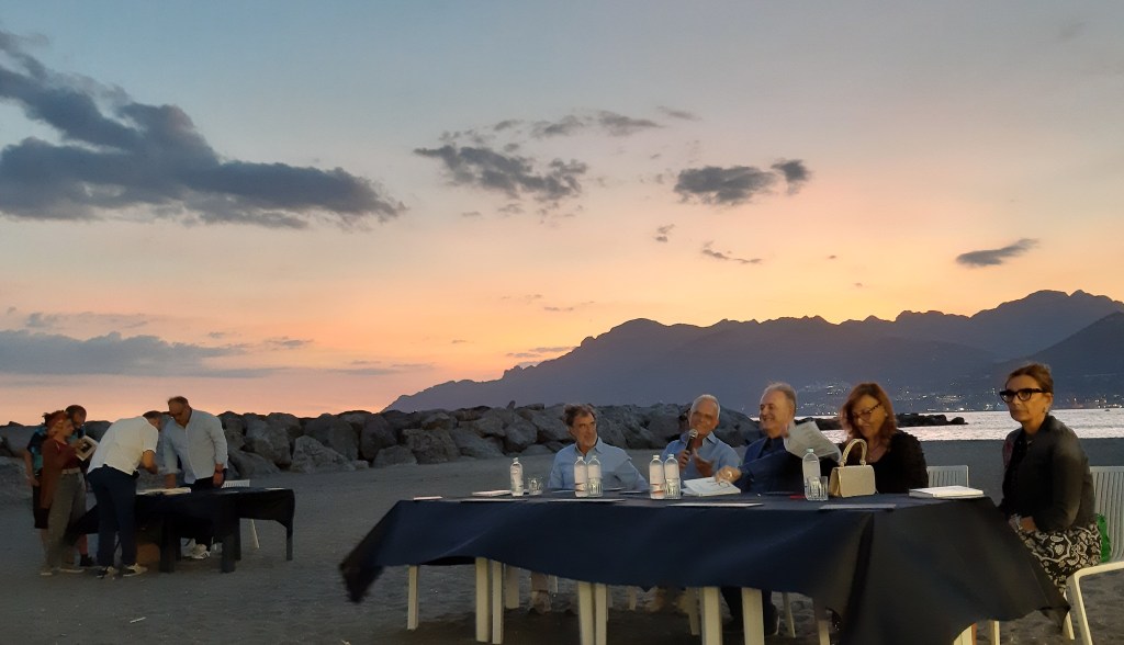 Un gruppo di persone sedute intorno a un tavolo all'aperto mentre il sole tramonta all'orizzonte, con le montagne sullo sfondo.