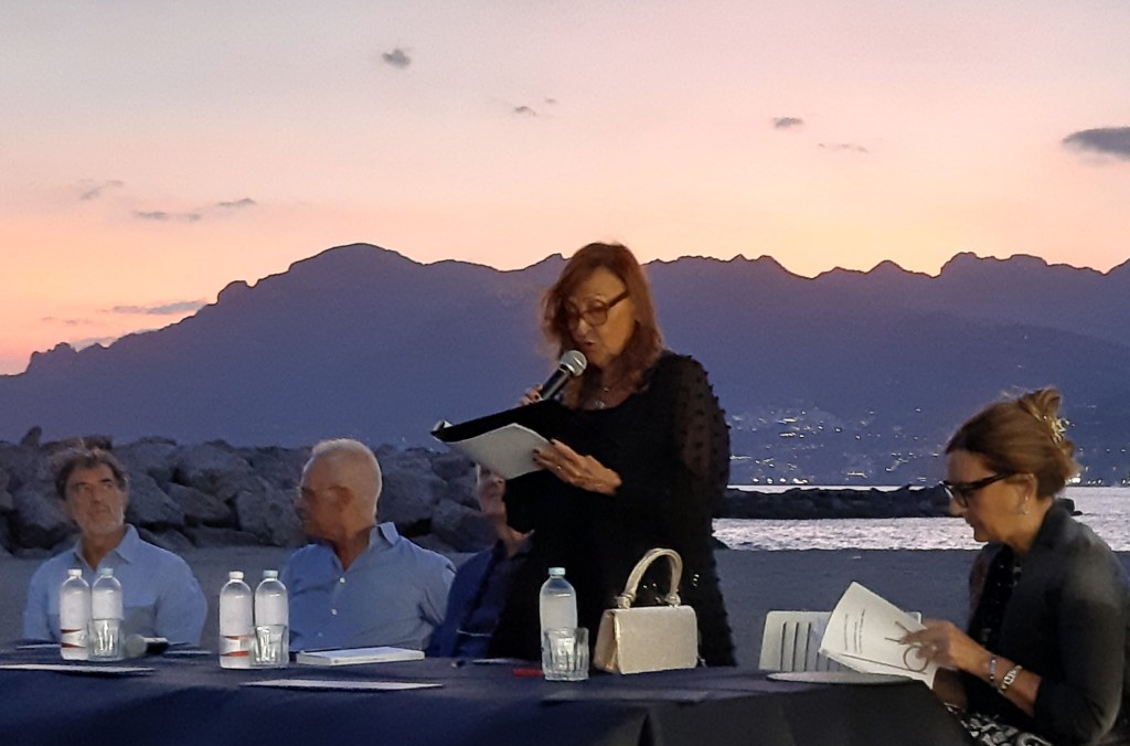 Una donna legge durante un evento all'aperto al Porticciolo di Pastena a Salerno, con un tramonto sullo sfondo e persone sedute attorno a un tavolo.