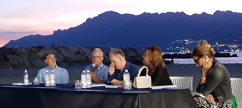 Un evento letterario al Porticciolo di Pastena a Salerno, con quattro relatori seduti a un tavolo, mentre il sole tramonta sullo sfondo montano. Atmosfera di condivisione e riflessione.