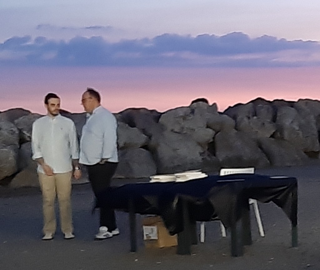 Due uomini conversano al Porticciolo di Pastena al tramonto, con rocce e un tavolo apparecchiato in background.