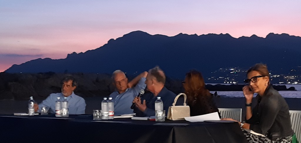 Un evento al Porticciolo di Pastena a Salerno, con relatori seduti a un tavolo, mentre il sole tramonta alle spalle, creando un'atmosfera suggestiva.