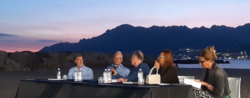 Un evento al Porticciolo di Pastena a Salerno, con un gruppo di relatori seduti a un tavolo, durante una serata al tramonto.