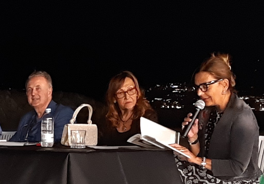 Un evento serale al Porticciolo di Pastena a Salerno, con un gruppo di relatori seduti attorno a un tavolo, mentre una donna legge da un libro. Sullo sfondo, si intravede l'illuminazione della città di Salerno.