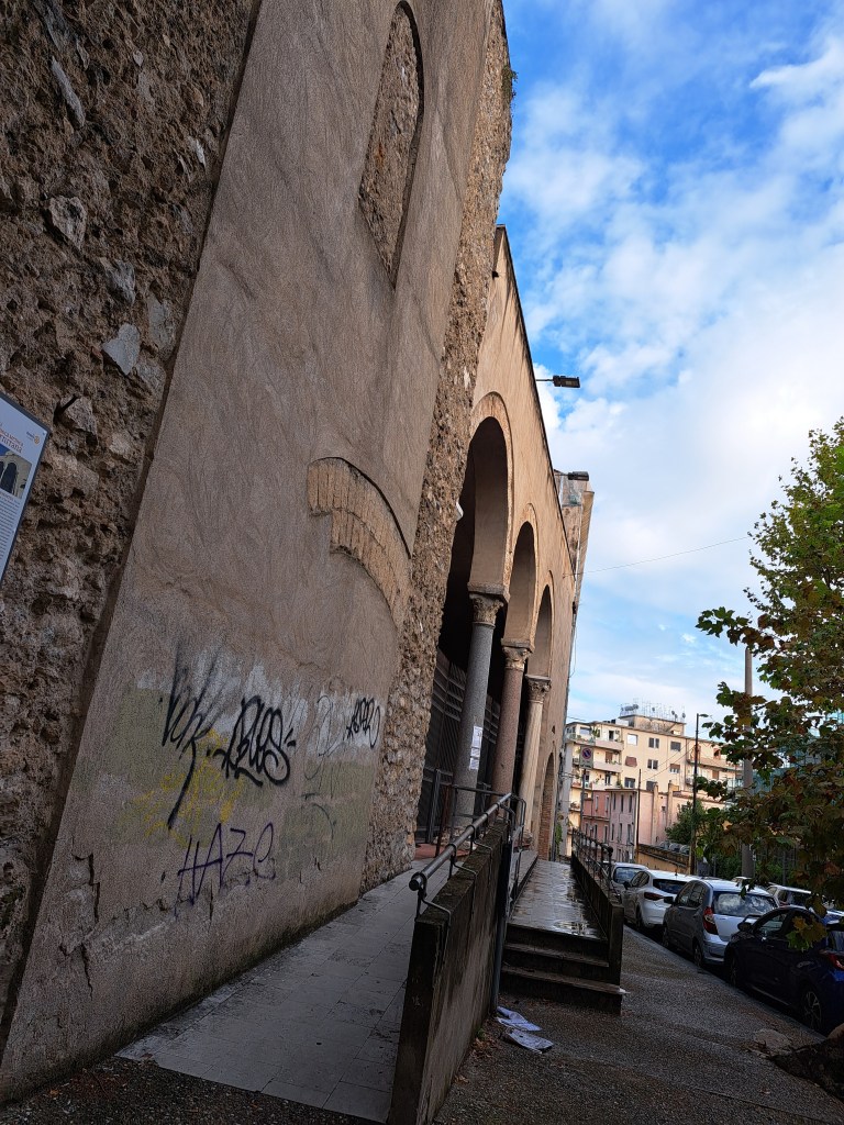 Facciata di un edificio storico con graffitti, vista inclinata lungo un marciapiede bagnato, con alberi e auto in secondo piano.