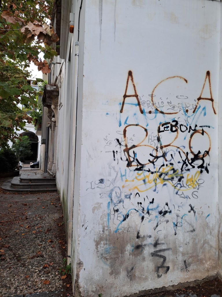 Murale con graffito su un muro bianco, con scritte e segni di vernice di diversi colori. Il terreno circostante è coperto di foglie secche, e si intravede una scala che porta verso un ingresso.