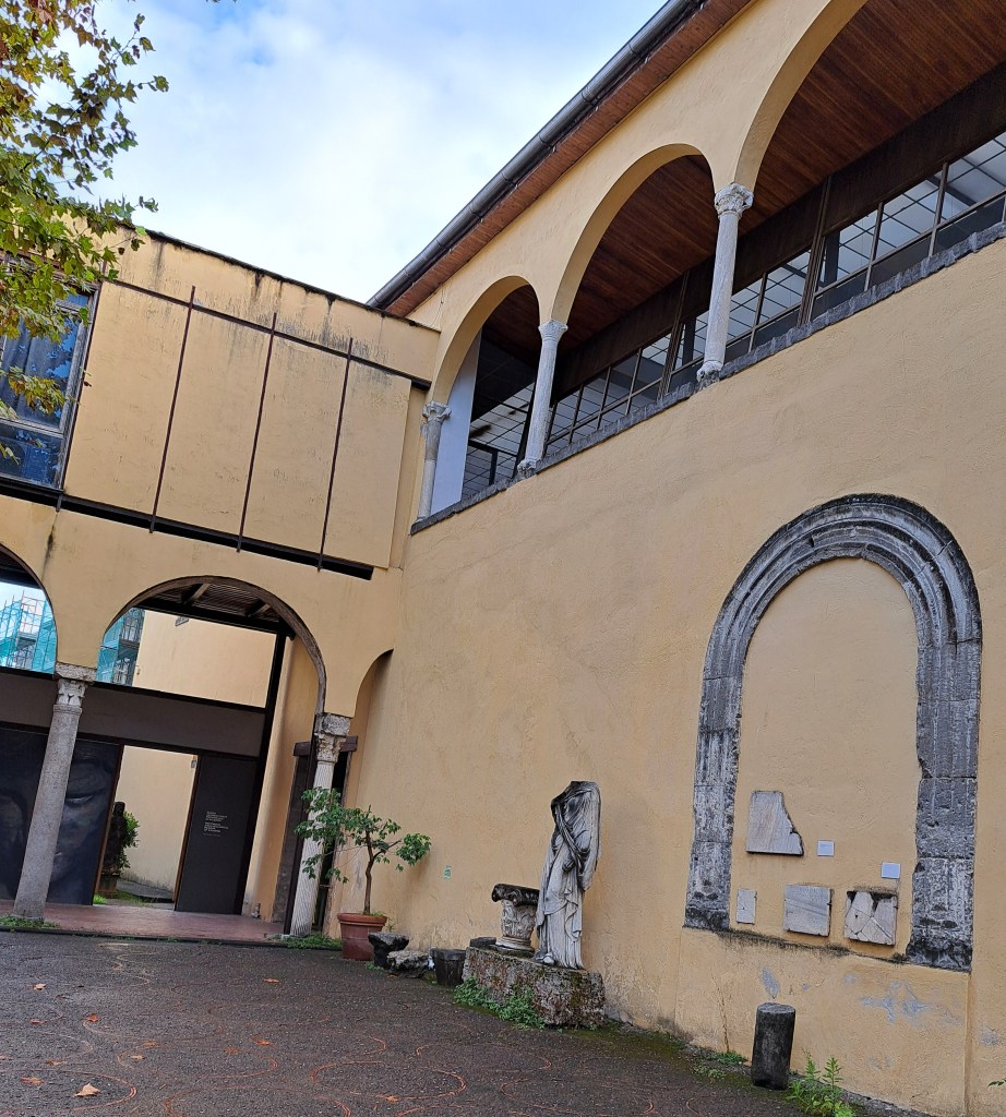 Vista di un cortile con un edificio di colore giallo, colonne e un'area di pavimentazione. Una scultura senza testa è visibile accanto a una parete che presenta tessere di diverso materiale. Un albero in un vaso è presente sul lato destro.