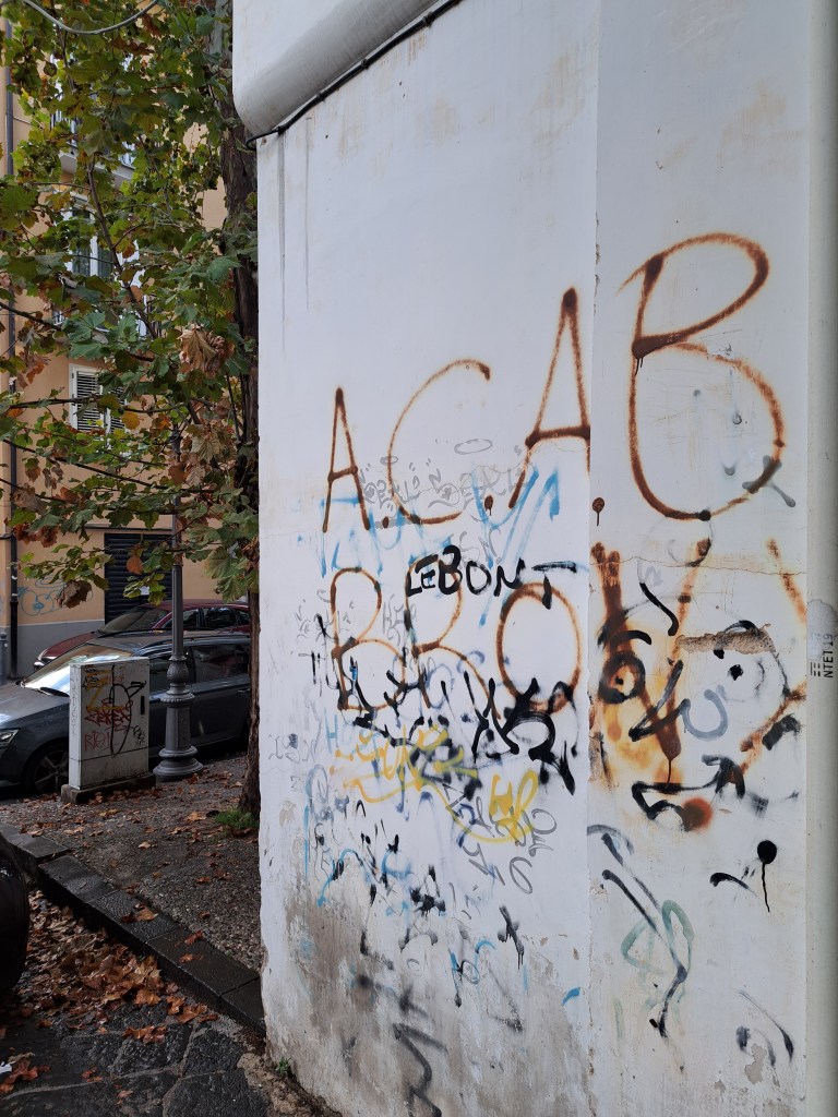 Una parete di un edificio con graffiti, inclusi scritte come 'ACAB' e 'BRC', circondata da alberi e un marciapiede.