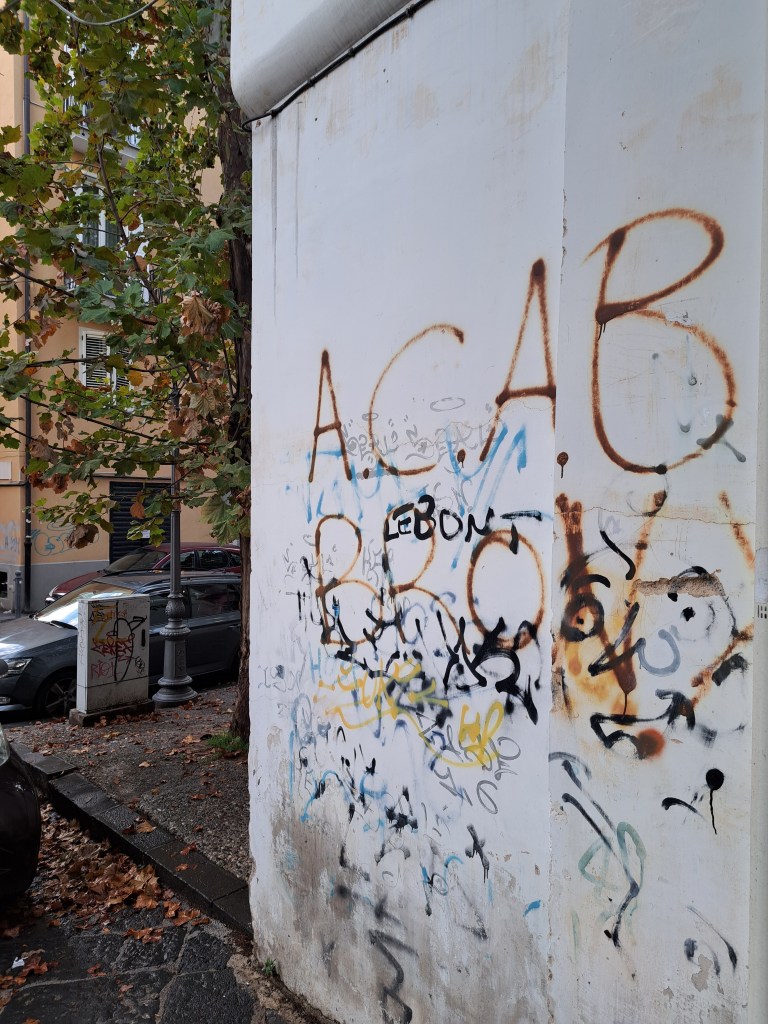 Muro di un edificio con graffiti colorati e inscripti, circondato da alberi e veicoli parcheggiati.