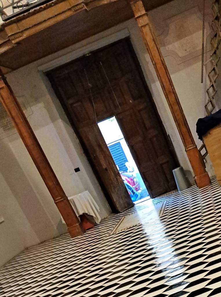 Interno di un edificio con una grande porta di legno aperta che dà su un esterno visibile, pavimento a motivi a scacchiera neri e bianchi, e una scala a vista.