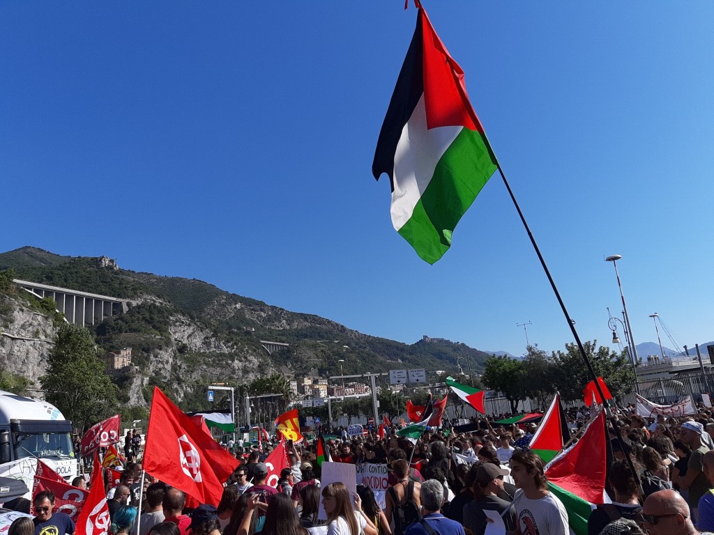Una grande manifestazione con persone che sventolano bandiere palestinesi e rosse, in un contesto urbano con montagne sullo sfondo e un cielo blu.