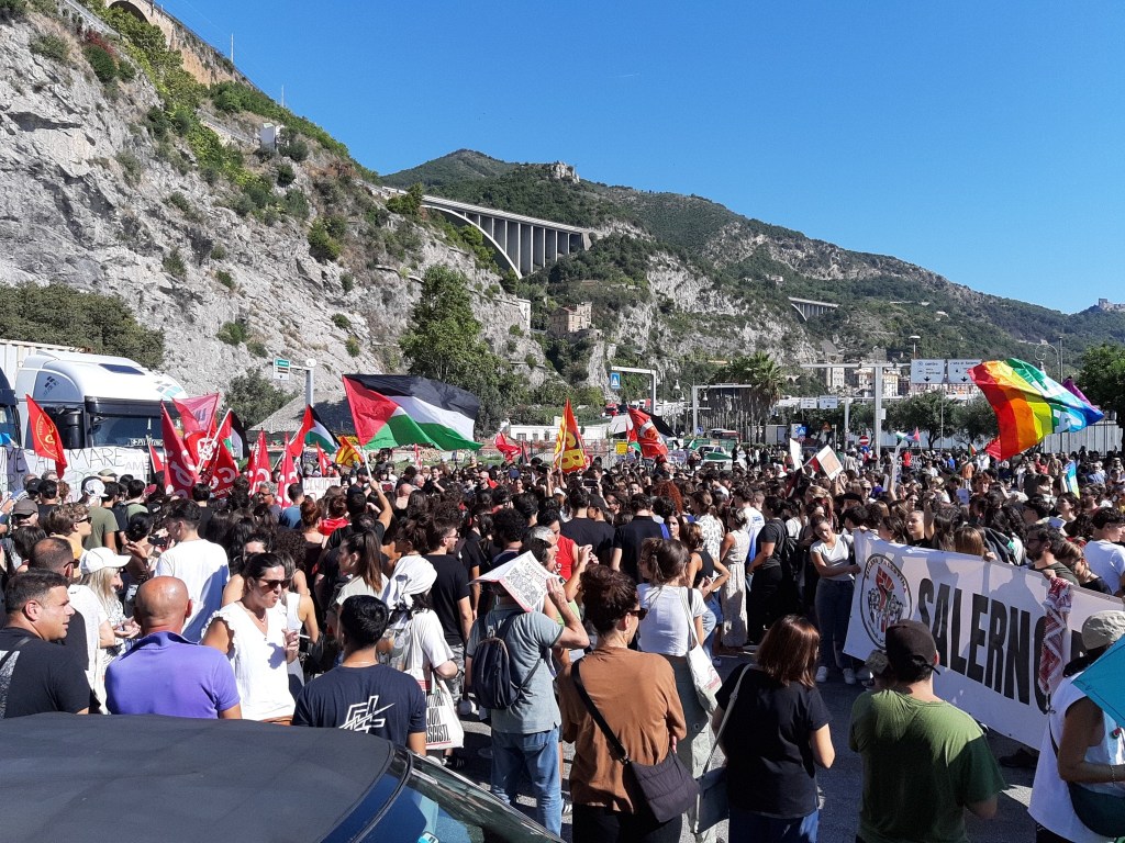 Una folla di manifestanti radunati sotto un viadotto, con diverse bandiere che sventolano, in un contesto di protesta.