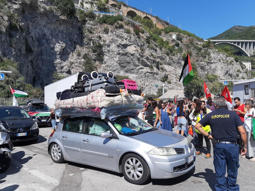 Una manifestazione di protesta con un'auto caricata di beni bloccata nel traffico, circondata da persone con bandiere e cartelli.