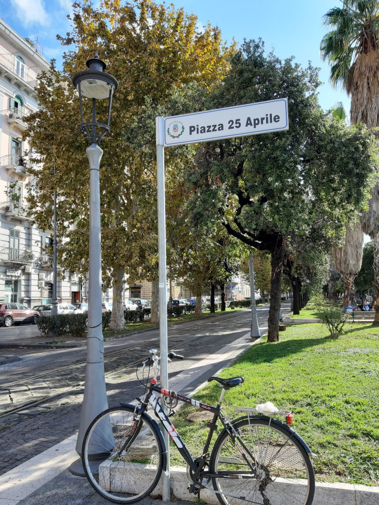 Un'insegna stradale che indica 'Piazza 25 Aprile' con una bicicletta parcheggiata accanto a un lampione, circondata da alberi in un'area verde, sotto un cielo sereno.