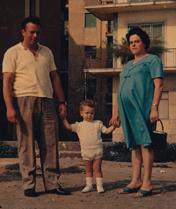 A family portrait featuring a man and a pregnant woman holding hands with a young child, standing outdoors in a residential area.