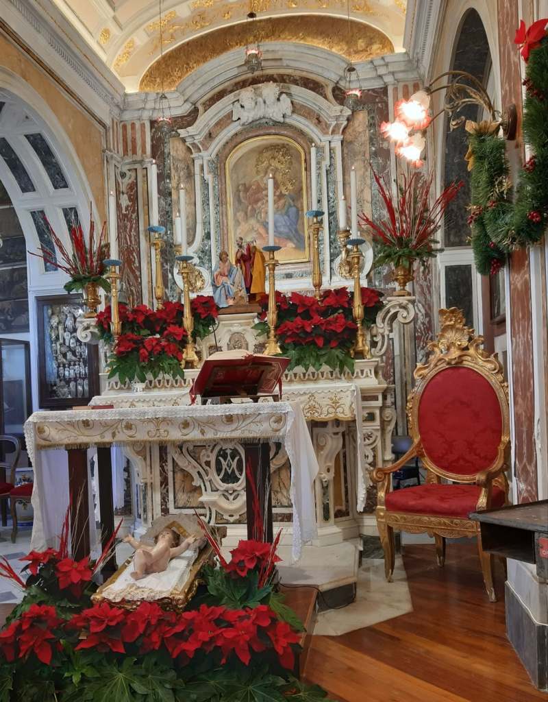 Interno di una chiesa con un altare decorato da fiori rossi, una statua del bambino Gesù in una culla dorata e una sedia di legno dorato, in un'atmosfera solenne.