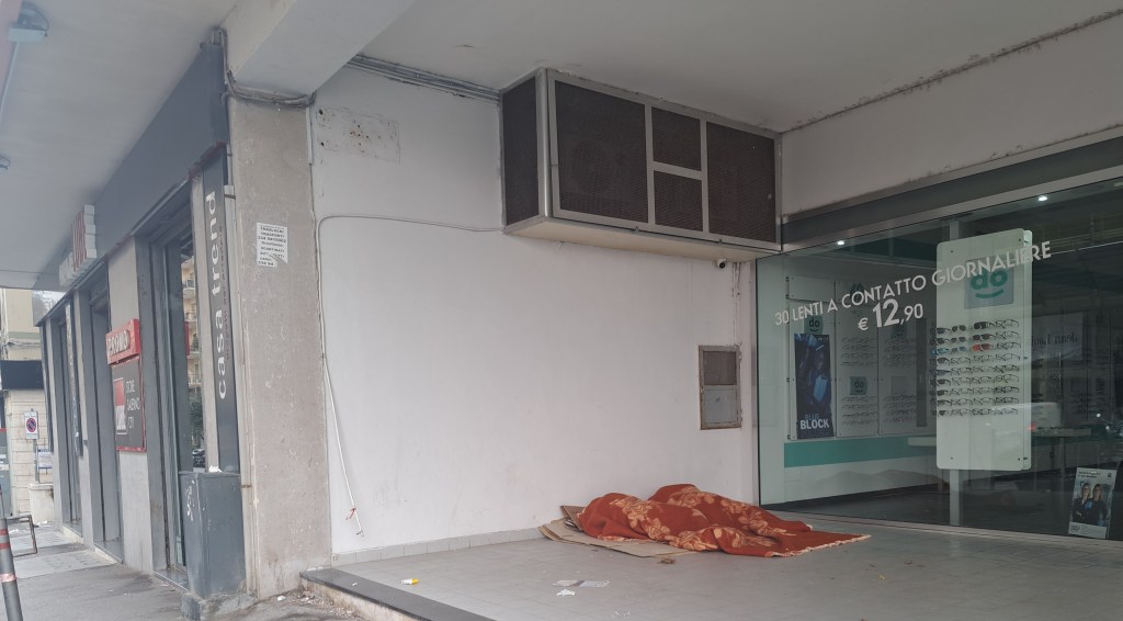 Un angolo di strada con un pavimento di piastrelle, un'affittanza di un negozio accanto e un tappeto arancione sul marciapiede.