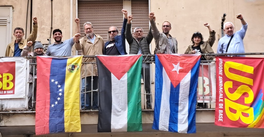 Un gruppo di persone solleva il pugno in segno di solidarietà su un balcone, circondato da bandiere del Venezuela, Palestina, Cuba e il banner USB.