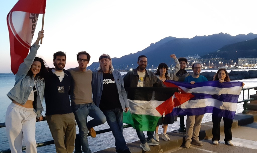Un gruppo di otto persone in piedi lungo una banchina, sorridendo e alzando bandiere. Sono presenti una bandiera palestinese e una cubana, con uno sfondo di mare e monti al tramonto.