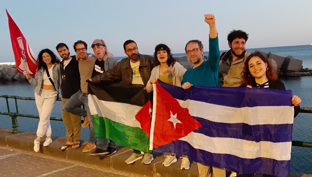 Gruppo di persone che posano insieme su un molo, ognuno con bandiere palestinesi e cubane, in un ambiente all'aperto al tramonto.