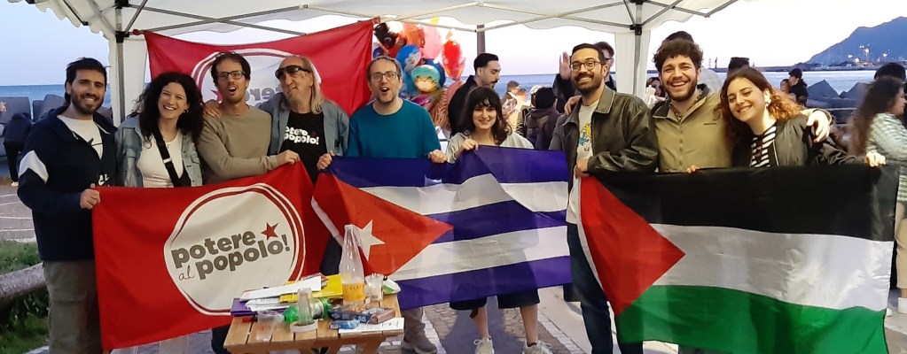 Un gruppo di persone in piedi sotto una tenda con bandiere di Cuba, Palestina e un manifesto di Potere al Popolo!.