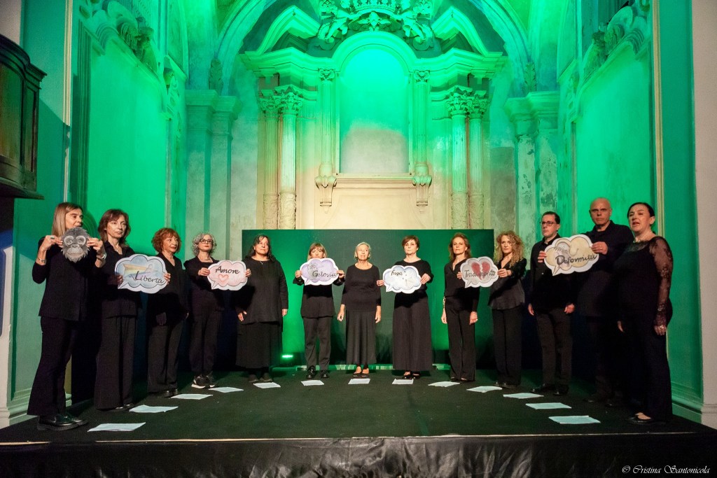 Un gruppo di persone in abiti neri sul palco, ognuna tiene in mano un cartello con parole significative come 'Libertà', 'Amore' e 'Gloria', illuminati da luci verdi.