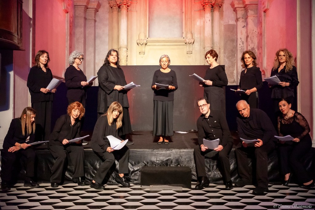 Gruppo di persone in abbigliamento nero che recitano su un palco, con un'atmosfera teatrale e illuminazione rossa.