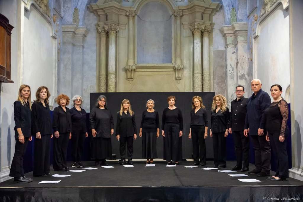 Un coro di donne e uomini in abbigliamento nero si esibisce su un palco in un ambiente architettonico elegante.