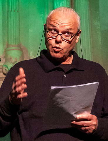 Un uomo con occhiali che legge appunti su un palco, mentre parla con espressione coinvolgente.