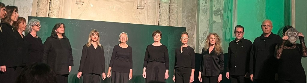 Gruppo di persone in abbigliamento nero durante una performance teatrale, con uno sfondo verde e dettagli architettonici decorati.
